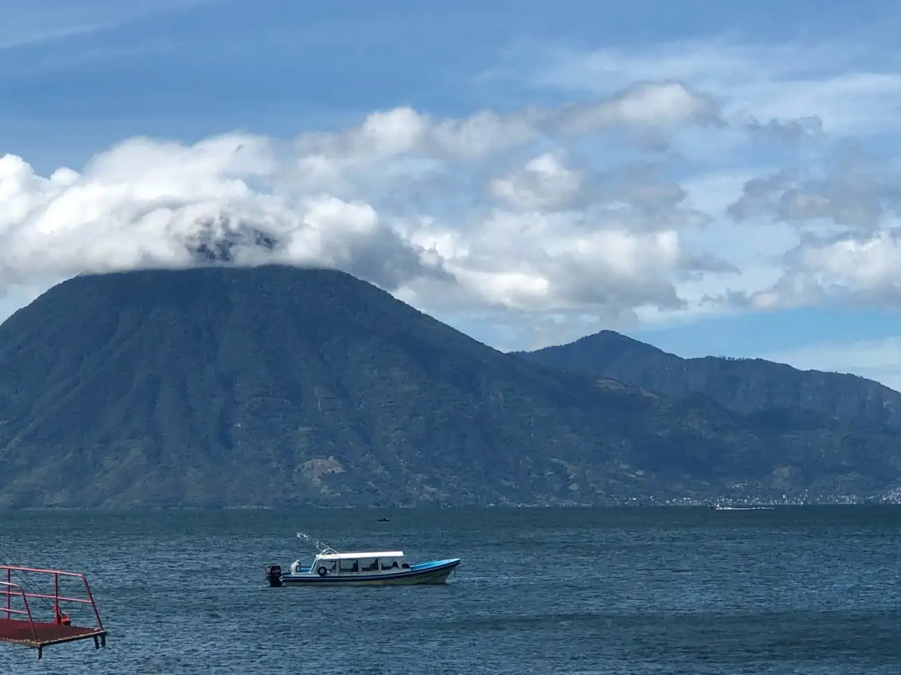 A body of water with a boat in the foreground and a mountain in the background with some clouds on the mountain's peak